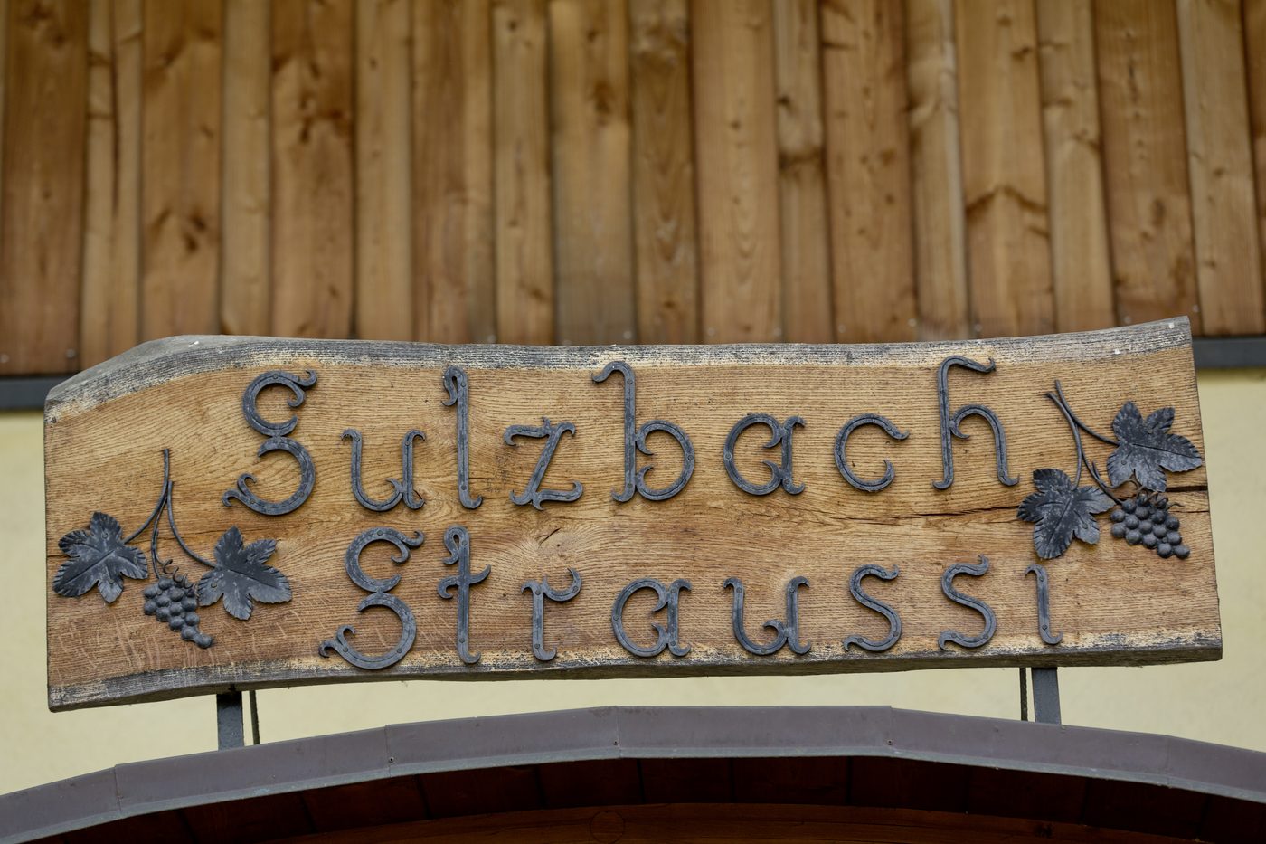 Handgeschmiedetes Holzschild „Sulzbach Straussi“ mit schmiedeeisernen Buchstaben und Rebendekor am Eingang
